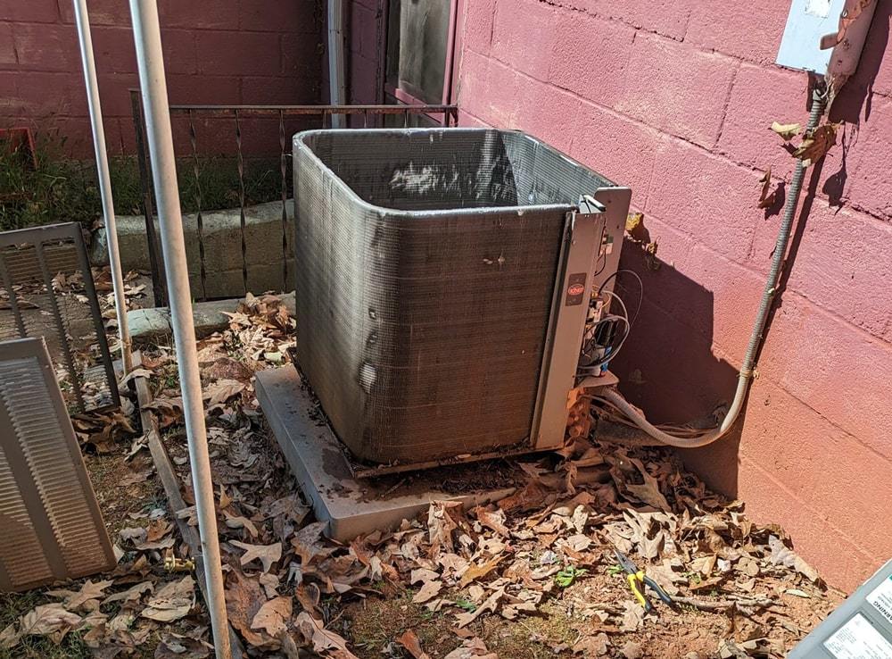 An old air conditioning unit sits on a concrete slab against a pink wall, surrounded by scattered dry leaves.