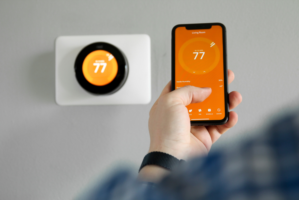 A person adjusts a smart thermostat via a smartphone app. Both displays show a temperature of 77 degrees.